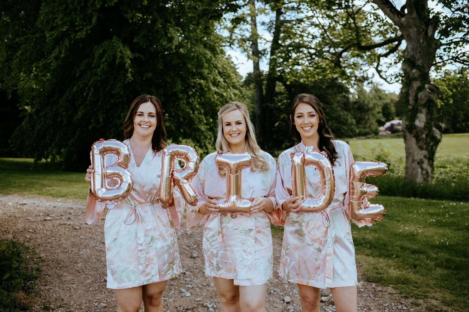 three bridesmaids standing outside wearing silk gowns and holding balloons that spell the word bride exclusive use colstoun house wedding unique wedding venue scotland