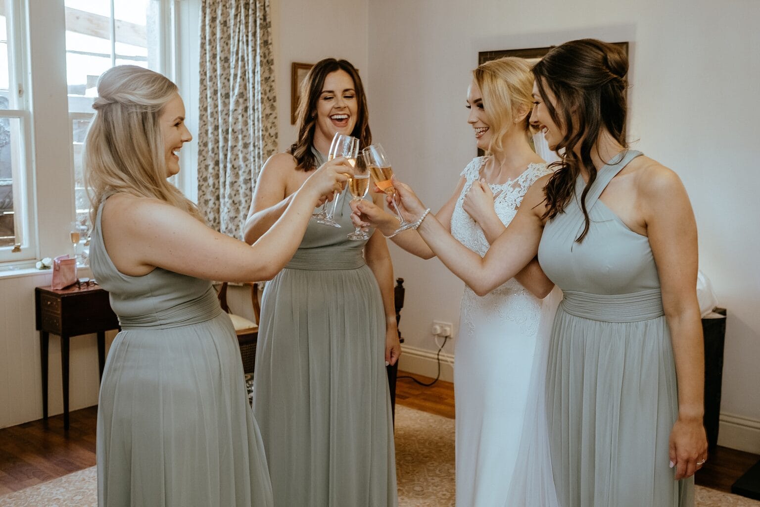 bride wearing white dress and three bridesmaids clinking glasses and smiling colstoun house wedding cost unique wedding venue scotland