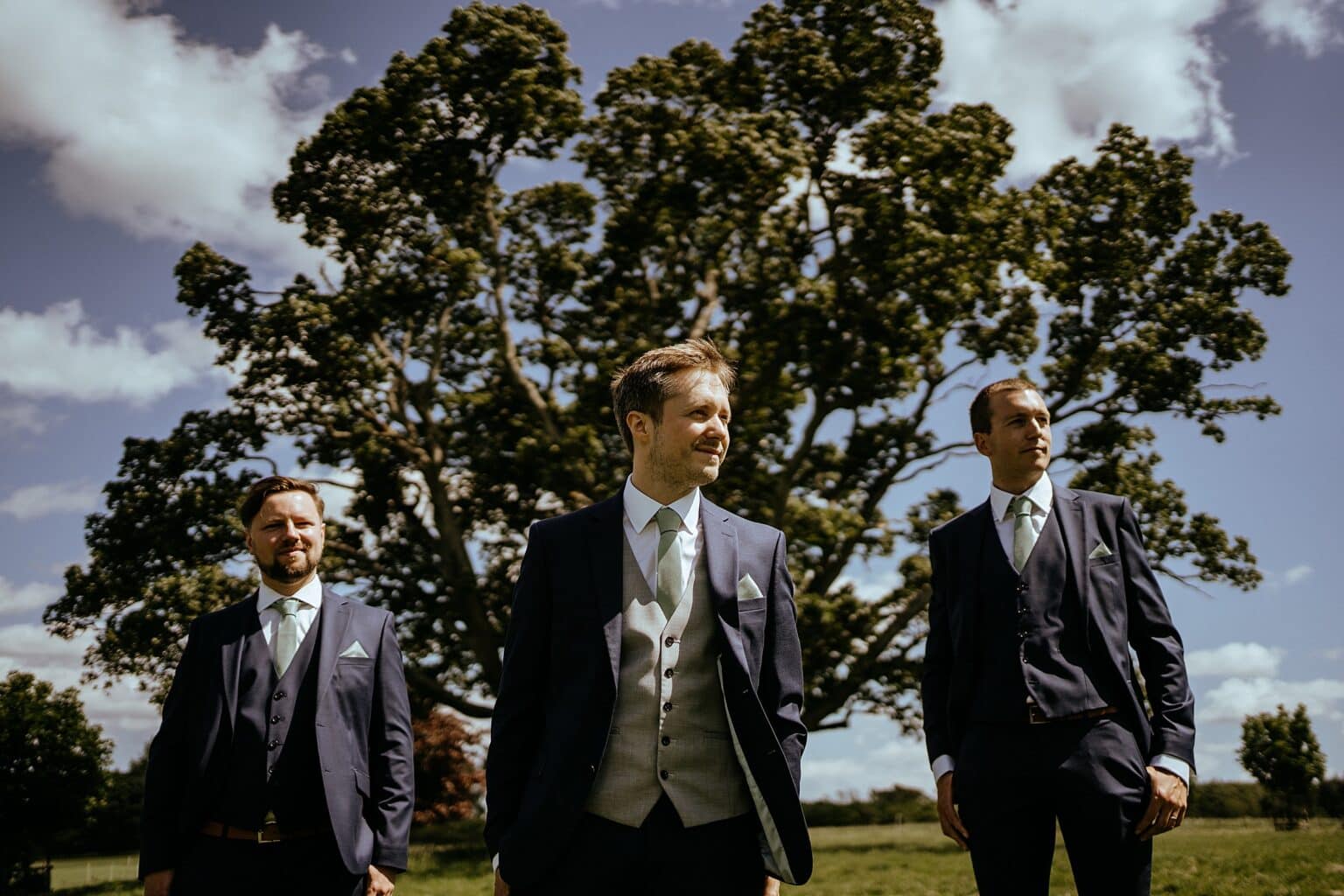 groomsmen standing to the side and behind the groom with big green tree and blue skies in background exclusive use colstoun house wedding unique wedding venue scotland
