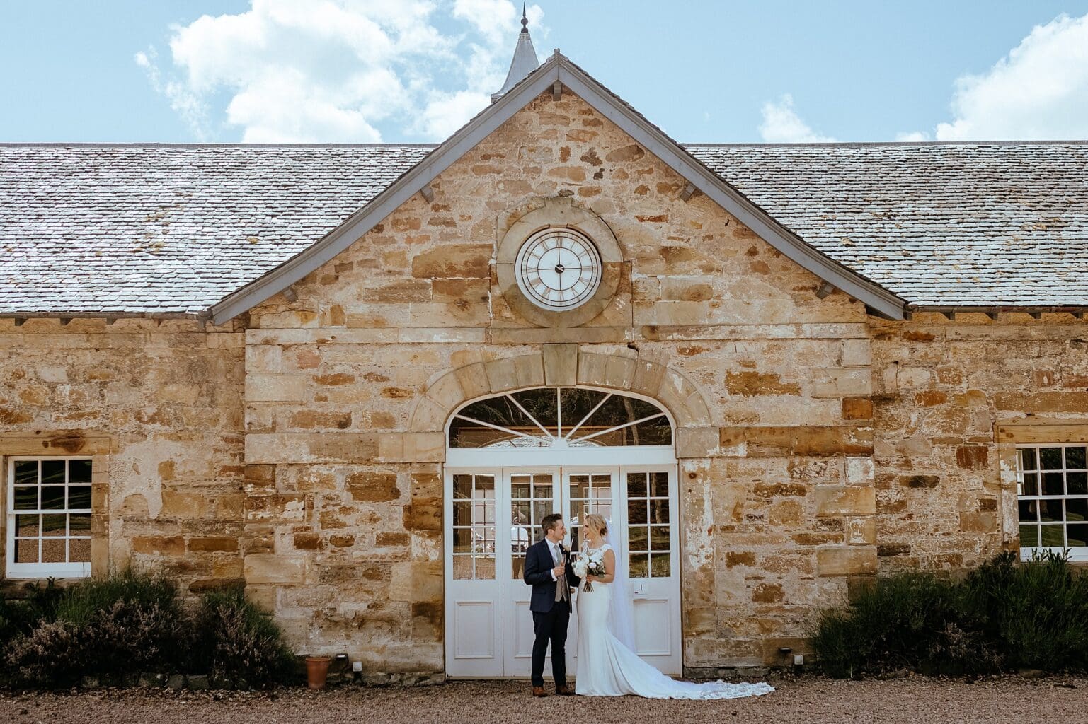 bride and groom standing in front of white door of stone building in courtyard on sunny day colstoun house wedding cost unique wedding venue scotland