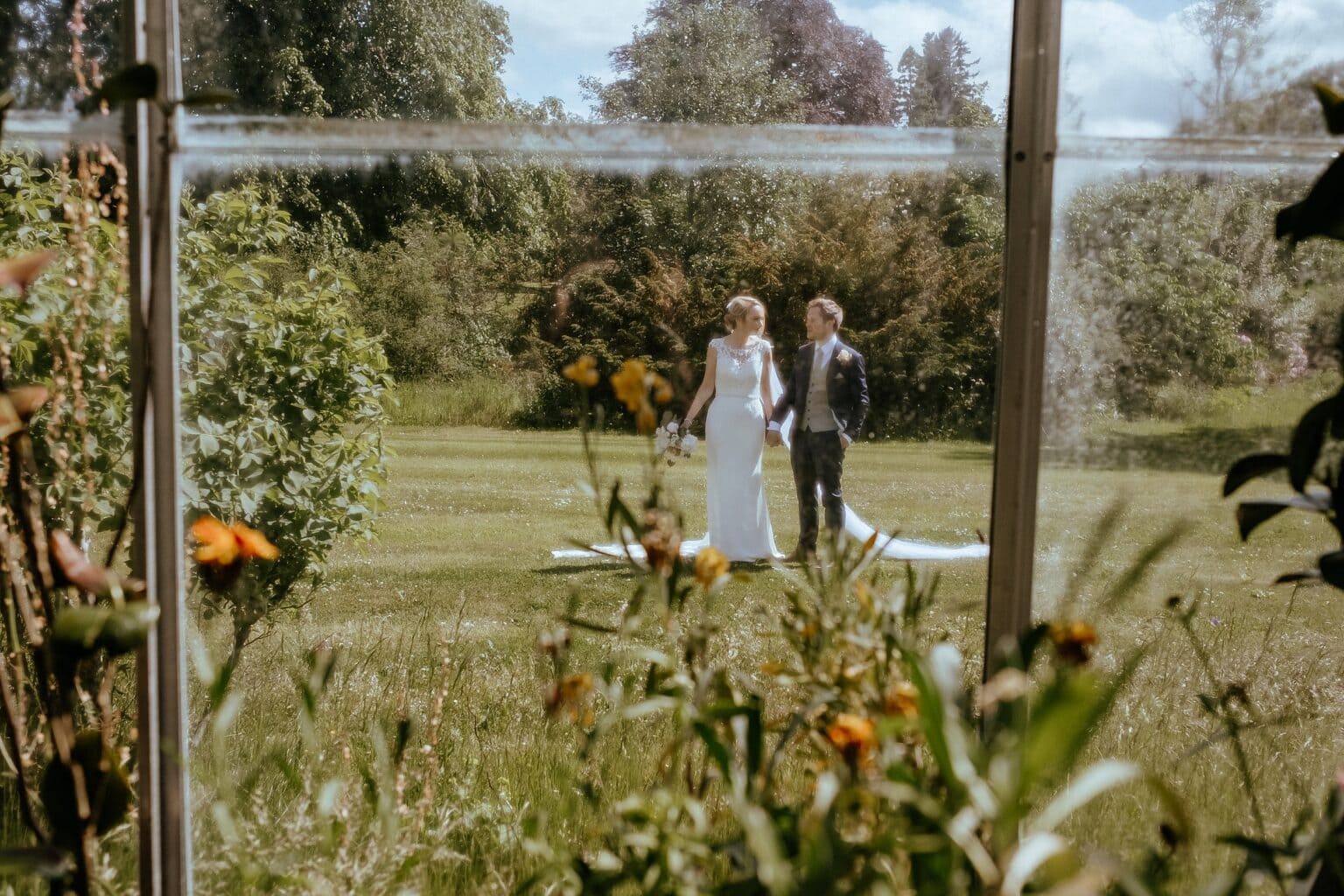 view from greenhouse of bride and groom walking through gardens on sunny day colstoun house wedding unique venues scotland unique wedding venue scotland