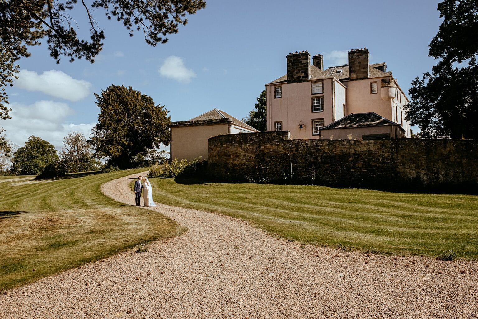 bride and groom walking up stone driveway of venue with grass lawns at each side and blue skies in background colstoun house wedding unique wedding venue scotland