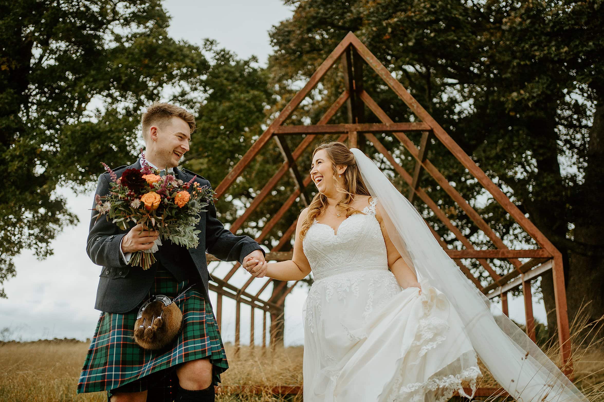 bride wearing white dress and groom wearing kilt outfit laughing as they hold hands in field with trees and building frame in background duntarvie castle wedding
