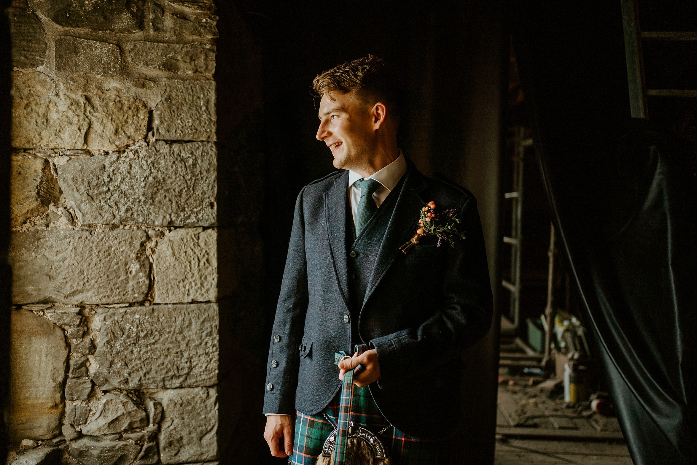 groom wearing kilt outfit in castle looking out of window as the light shines on his face duntarvie castle wedding