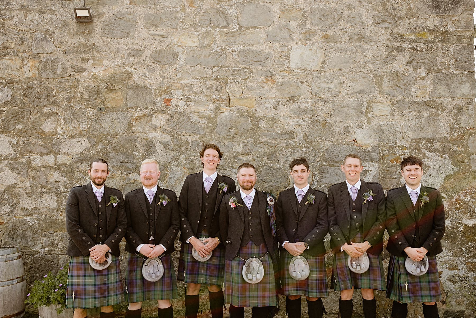 groom and groomsmen dressed in kilts standing in front of stone wall at the den at culross after their Scottish barn wedding