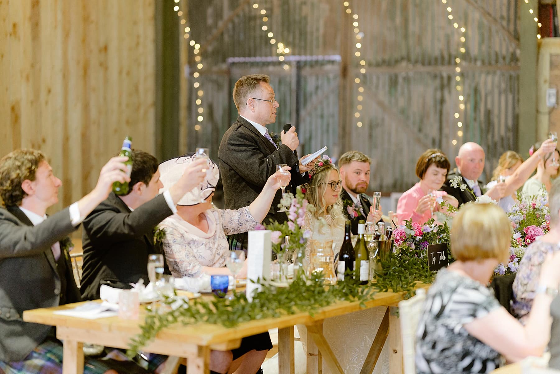 father of the bride giving speech inside barn in front of top table with green foliage and fresh flowers inside barn at the den at culross wedding