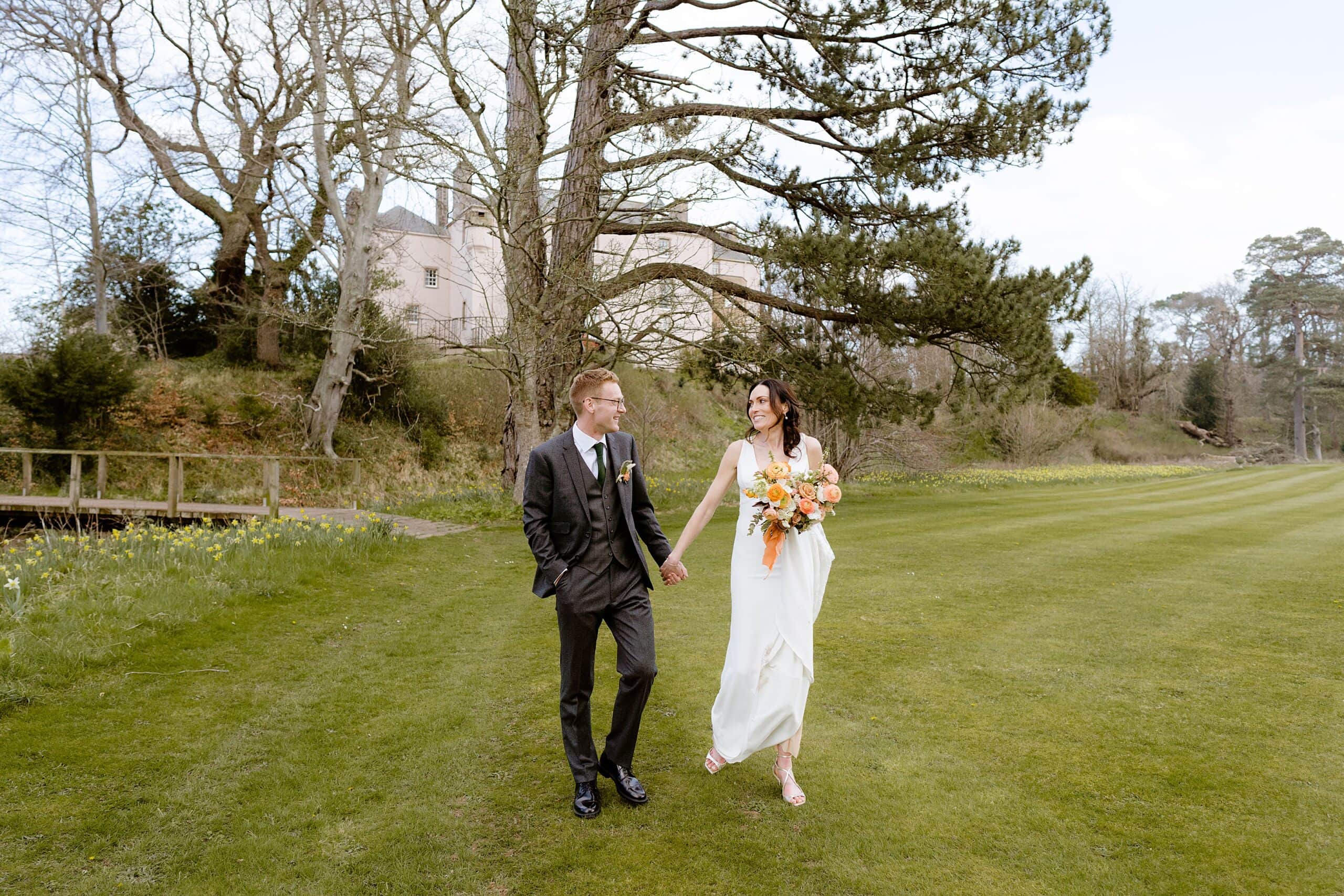 the bride and groom hold hands and smile at each other as they walk on a lawn with trees and a farm building in the background following their unusual wedding venues east lothian wedding ceremony near edinburgh scotland