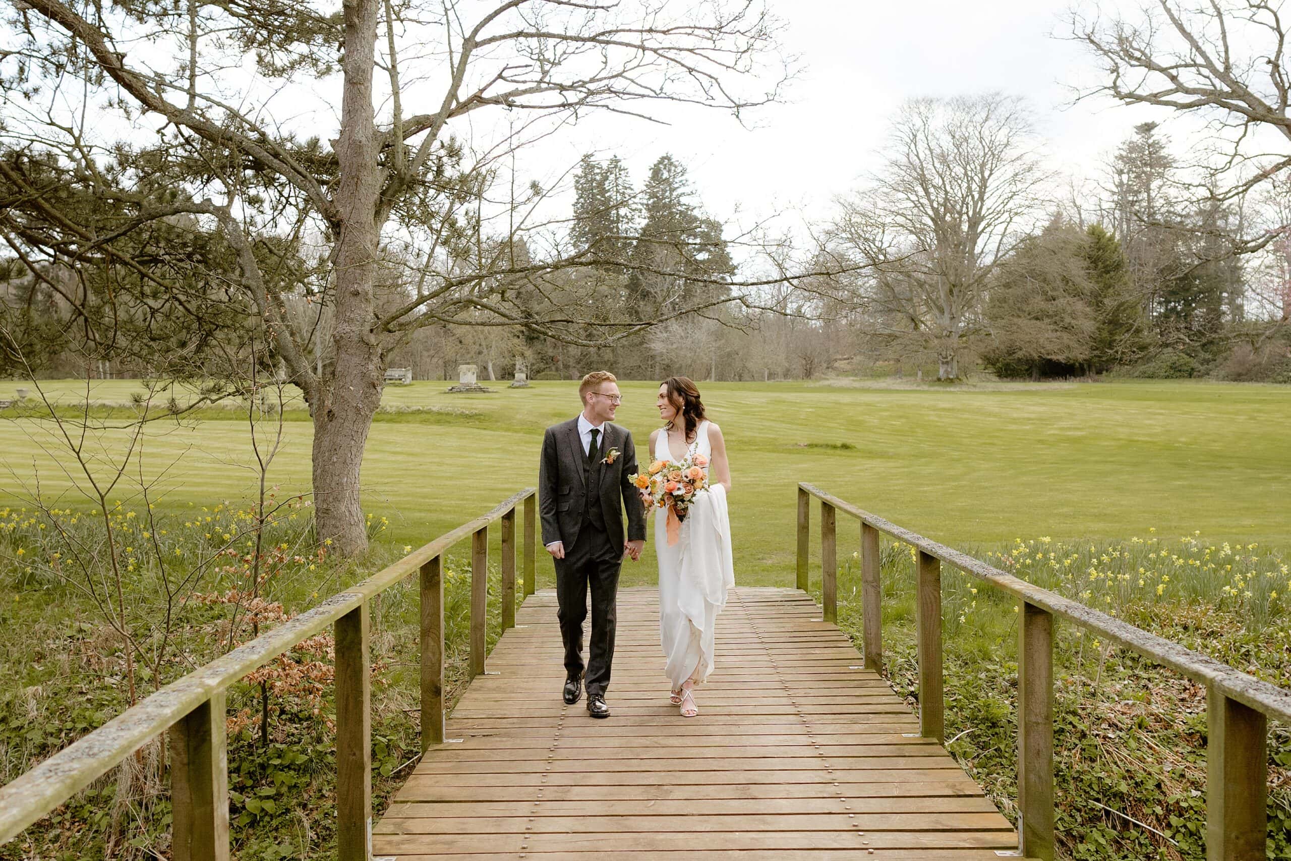 the bride and groom walking across a wooden bridge with lawn and trees in the background following their unusual wedding venues east lothian wedding ceremony near edinburgh scotland