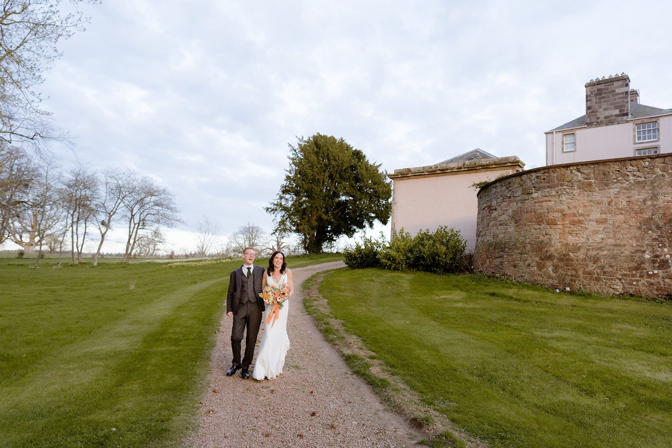 the bride and groom walking down a stone path with lawn trees and a farm building in the background following their unusual wedding venues east lothian wedding ceremony near edinburgh scotland