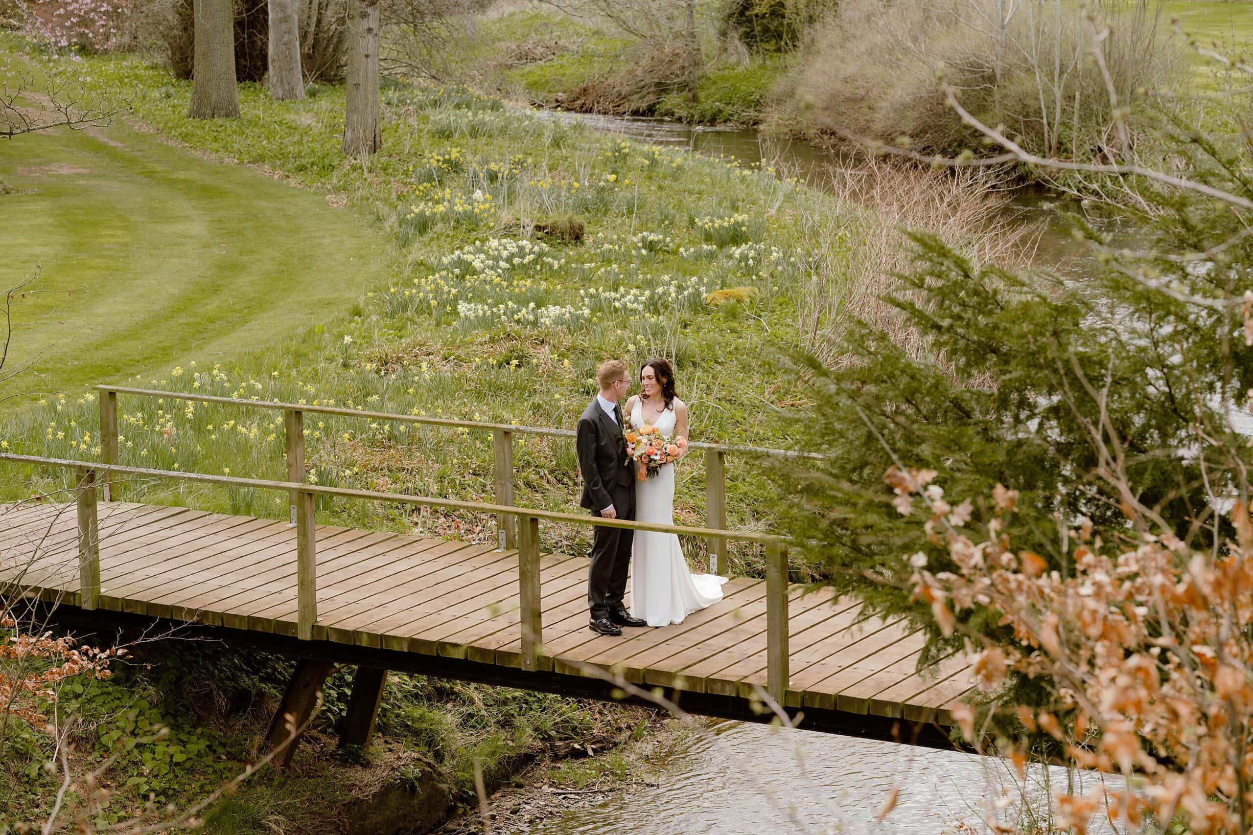 the bride and groom standing on a wooden bridge over a stream with lawn and trees in the background following their unusual wedding venues east lothian wedding ceremony near edinburgh scotland