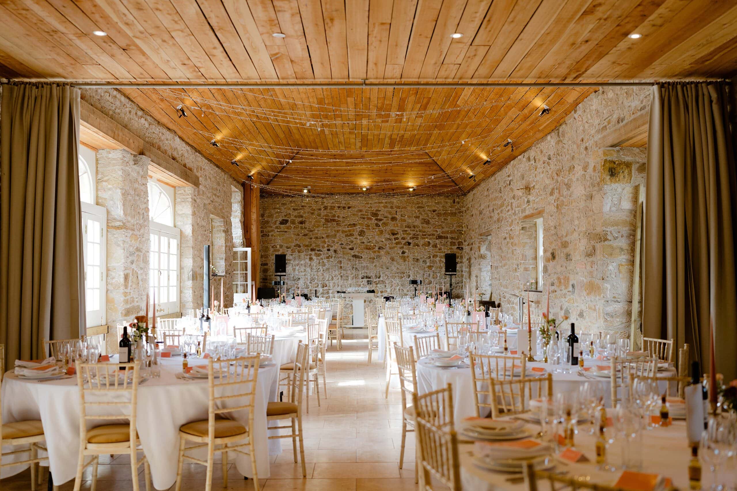 interior inside view of a farm wedding venue with stone walls and a wooden ceiling showing tables set for dinner and fairy lights hanging above