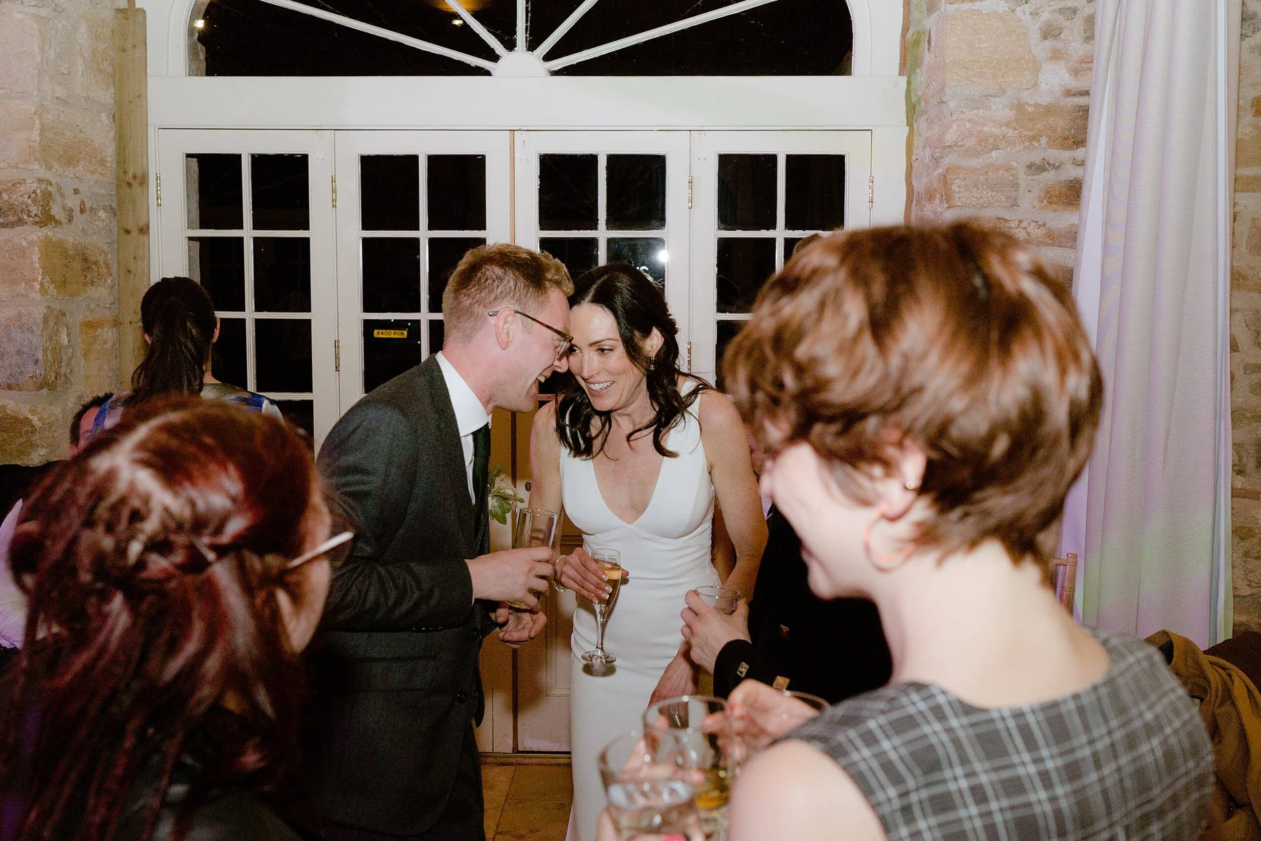 documentary shot of the bride and groom holding champagne glasses and leaning in towards each other at their relaxed wedding reception at their farm wedding venue in east lothian scotland