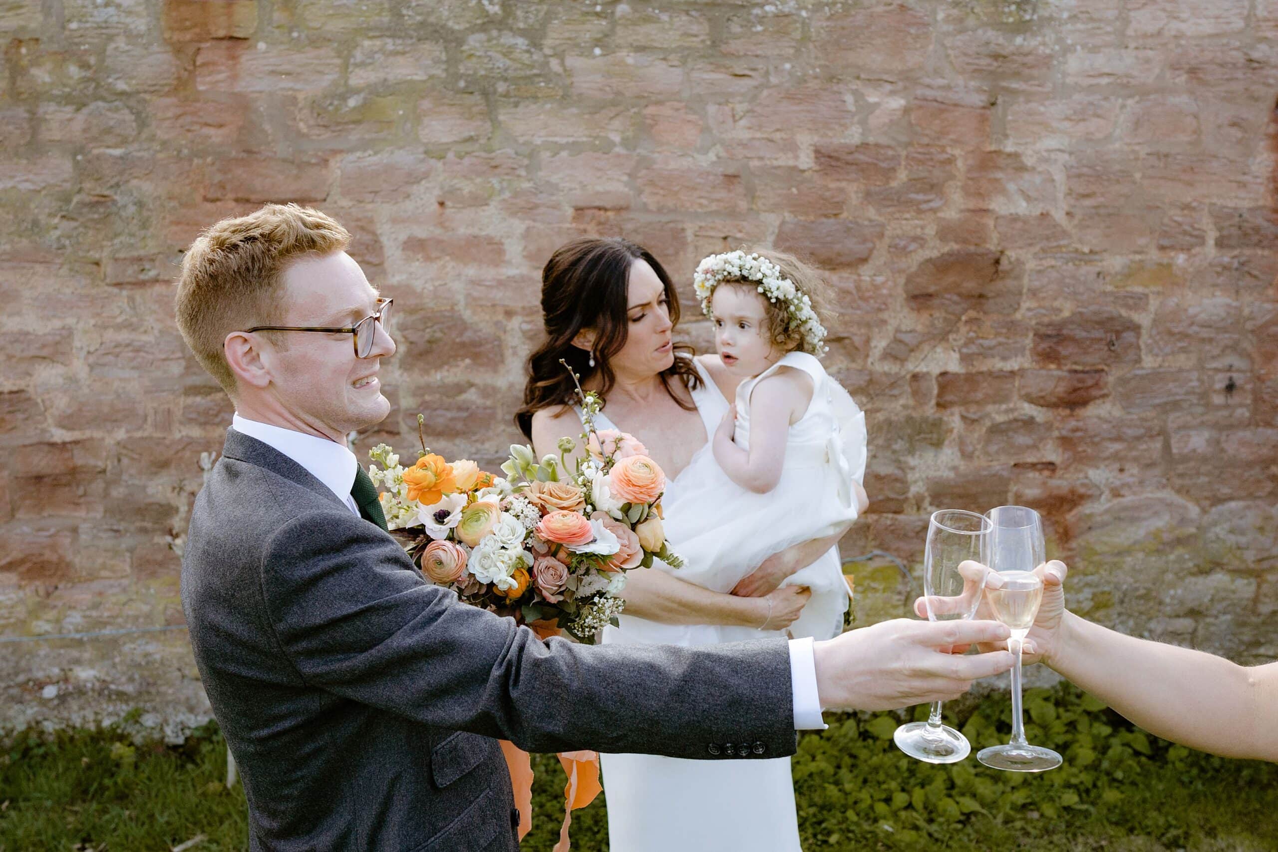 documentary shot of the bride holding her toddler daughter as the groom gives away a champagne glass as they stand in front of a stone wall at their outdoor wedding venue near edinburgh in scotland