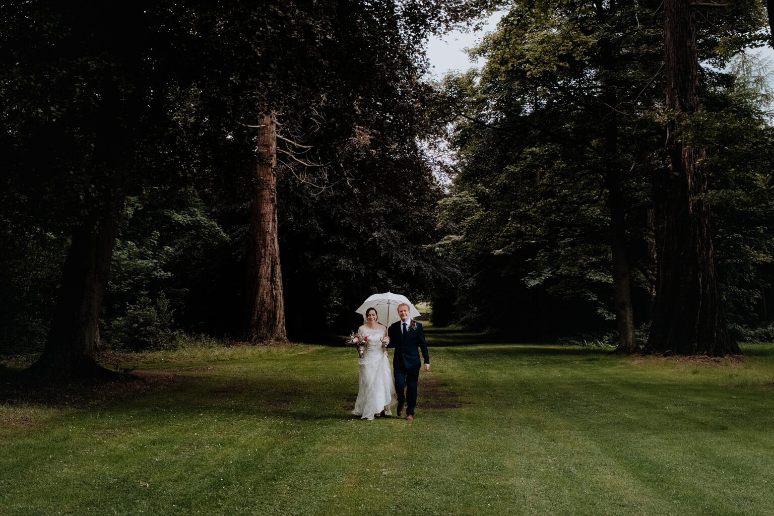 bride and groom walking towards camera across lawn with white umbrella with big trees in background what to do if it rains on your wedding day