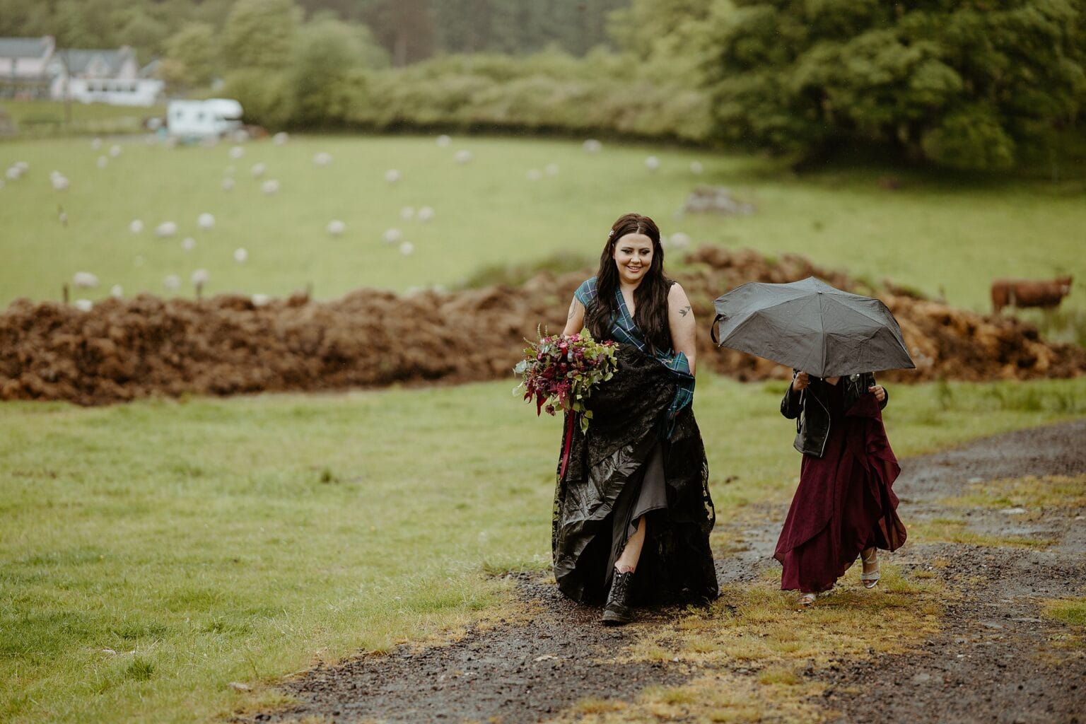 bride wearing black dress and black boots walking along country path in the rain as daughter holds umbrella what to do if it rains on your wedding day scottish wedding photography