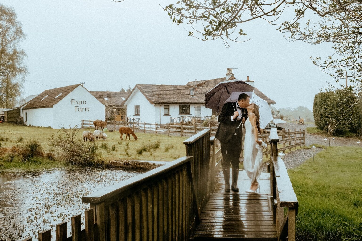 bride and groom standing on small wooden bridge kissing under umbrella on rainy day what to do if it rains on your wedding day